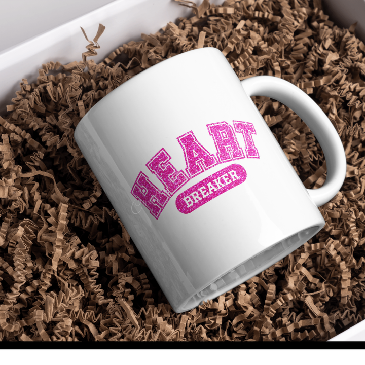 White ceramic mug featuring the ‘Heartbreaker’ design in bold pink glitter varsity-style text, placed on a bed of brown crinkle paper