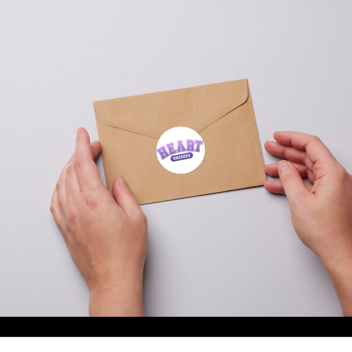 Hands holding a kraft paper envelope sealed with a circular sticker featuring the ‘Heartbreaker’ design in purple varsity-style text. The design adds a playful, custom touch to the envelope.