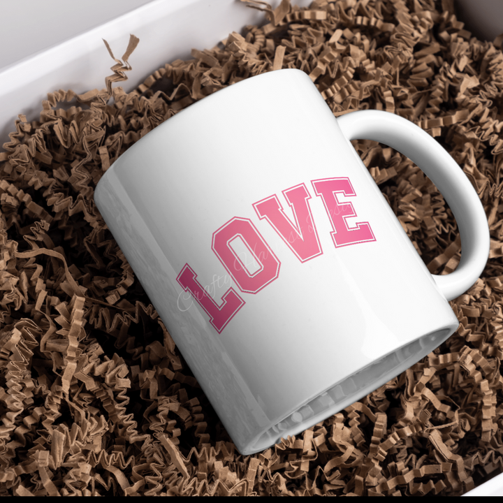 White ceramic mug featuring the ‘Love’ design in pink varsity-style text, placed on a bed of crinkle paper.