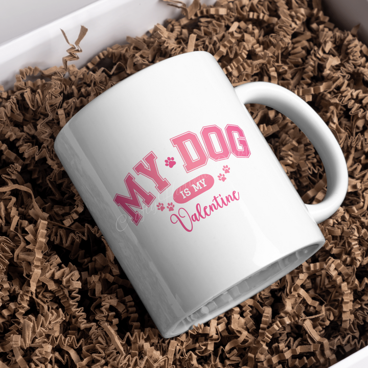 White ceramic mug featuring the ‘My Dog Is My Valentine’ design in pink varsity-style text with paw print accents, placed on a bed of crinkle paper.