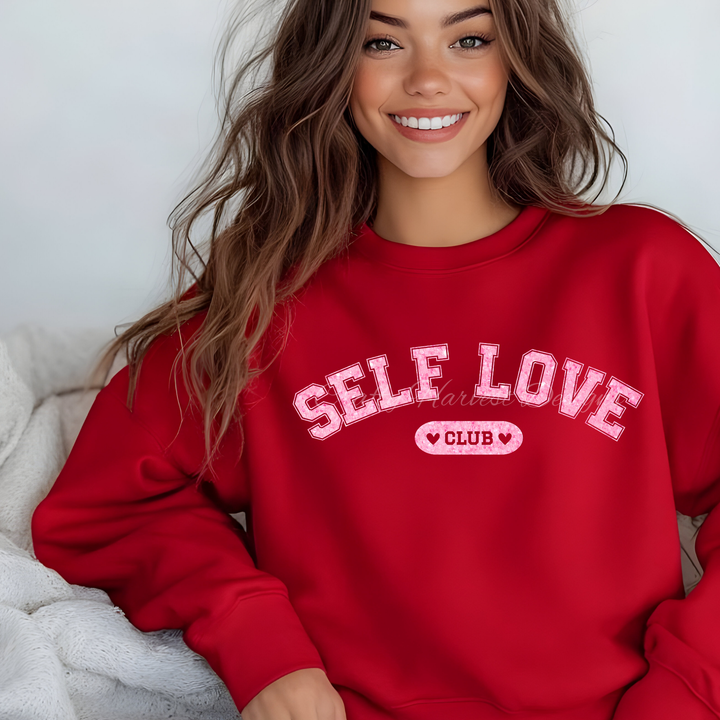 Model wearing a red sweatshirt featuring the ‘Self Love Club’ design in pink glitter varsity-style text with heart accents.