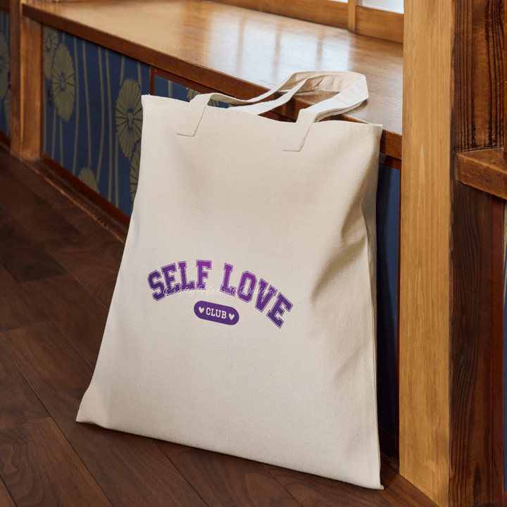 Beige tote bag featuring the ‘Self Love Club’ design in purple varsity-style text with heart accents.