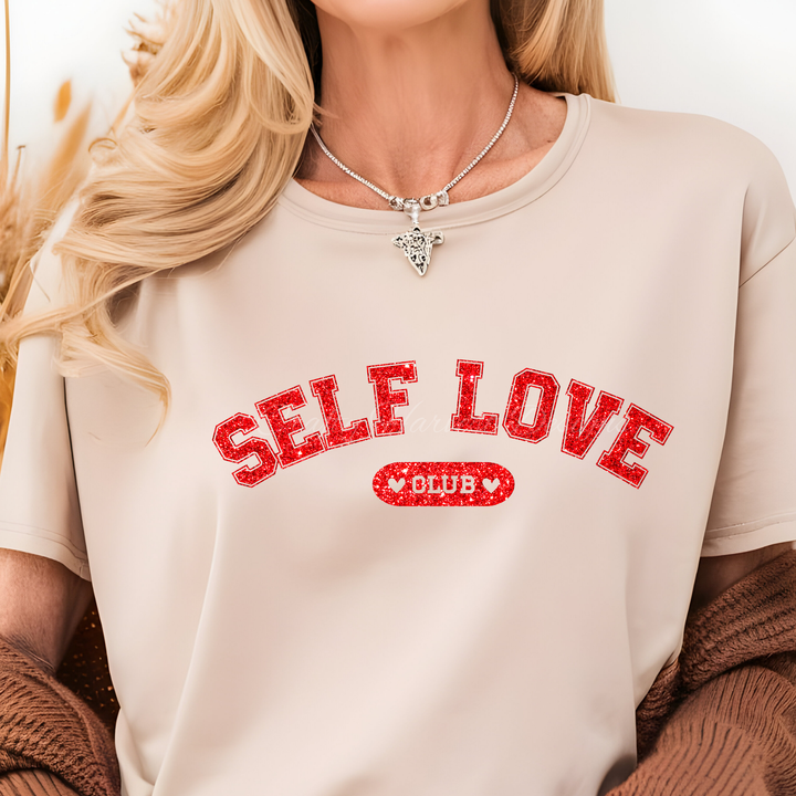 Model wearing a beige t-shirt featuring the ‘Self Love Club’ design in red glitter varsity-style text with heart accents.