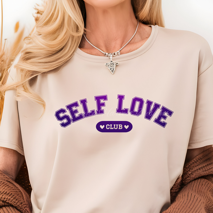 Beige t-shirt featuring the ‘Self Love Club’ design in purple varsity-style text with heart accents.